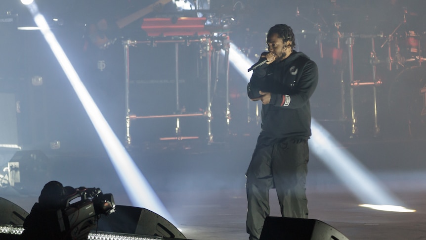 Kendrick Lamar, dressed in all black, raps on a big outdoor stage.