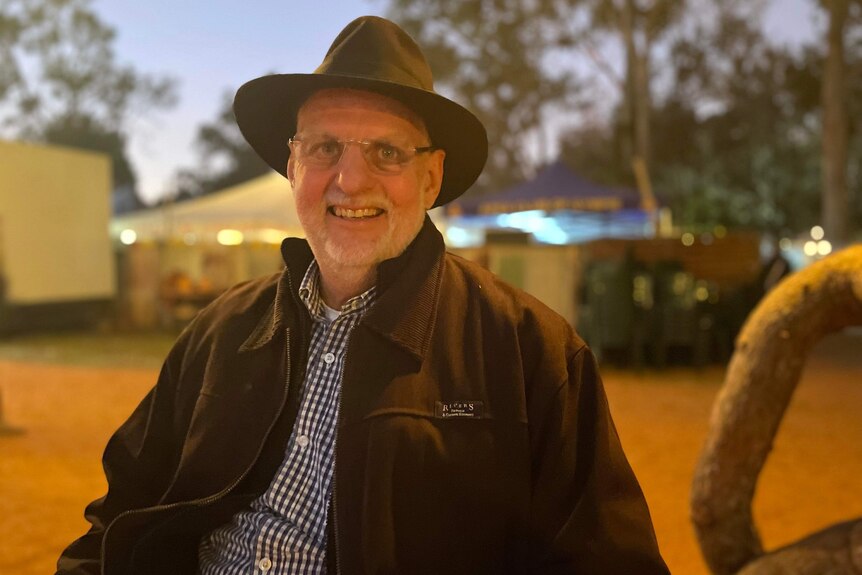 A man in a coat and a cowboy had smiles, there are tents behind him 