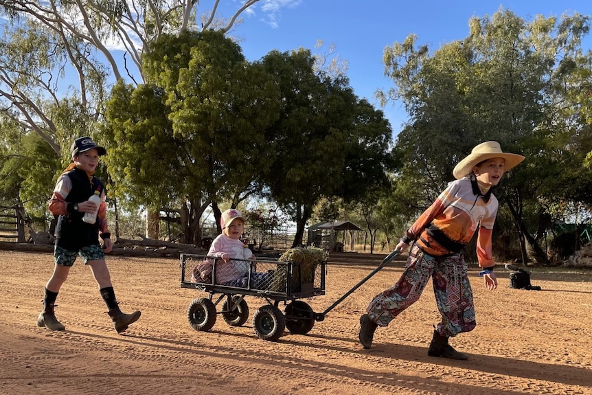 A child sits inside a trolly and is pulled along by another child along a dirt track