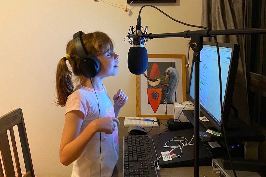 Young girl with headphones on talking into microphone in front of a computer.