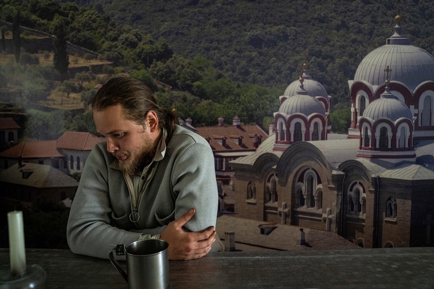 A man with a beard and long hair sits looking down at a table. A murakl of a city is painted behind him. 