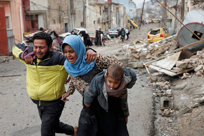 A man and a woman carrying a young child run, crying, through the ruined streets of Mosul.