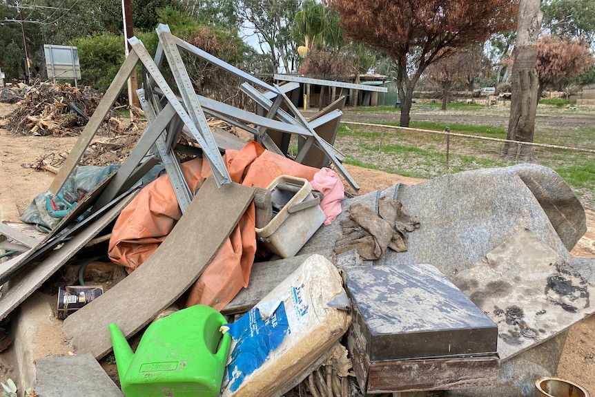 A pile of rubbish with assorted household items sits next to a road with a park on one side behind a short fence. 