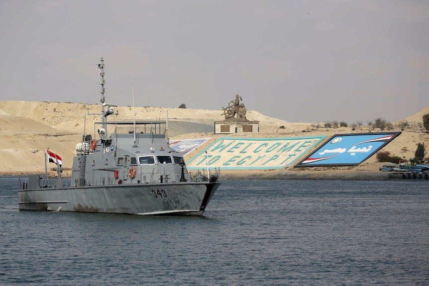 A grey water authority boat sails past a sandy bank. On the bank lies a sign that reads 'welcome to egypt'