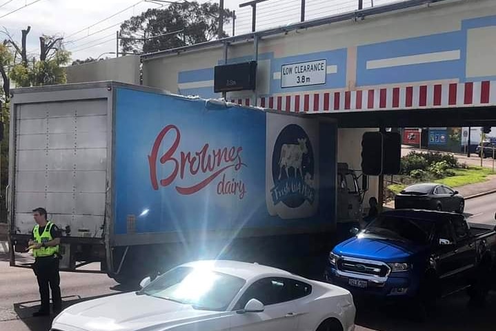 A Brownes Dairy truck is stuck under a bridge, with a police officer standing beside.