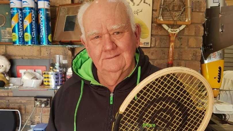 man balding with white hair in a black, grey and green hoodie holding a tennis racquet 