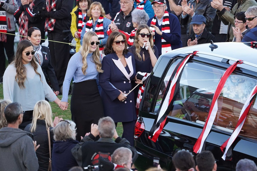 Four women holding walk behind a hearse draped with St Kilda football club flags.