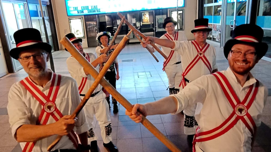 Six dancers in white outfits and top hats cross wooden sticks with each other while smiling