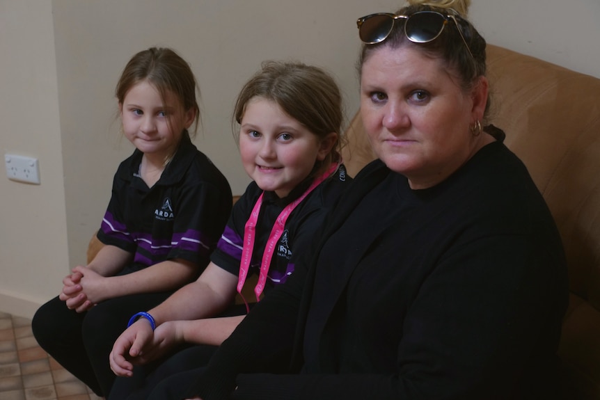 A mother and two young daughters sit on a brown couch.