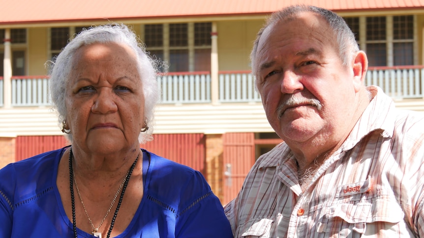 An elderly south sea islander woman sits next to an elderly white man. They look upset 