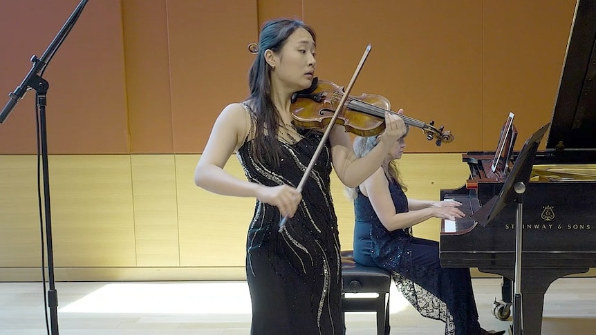 Yebin Yoo performs violin in front of her accompanist on a grand piano with an emotional expression, in a sparkly black dress.