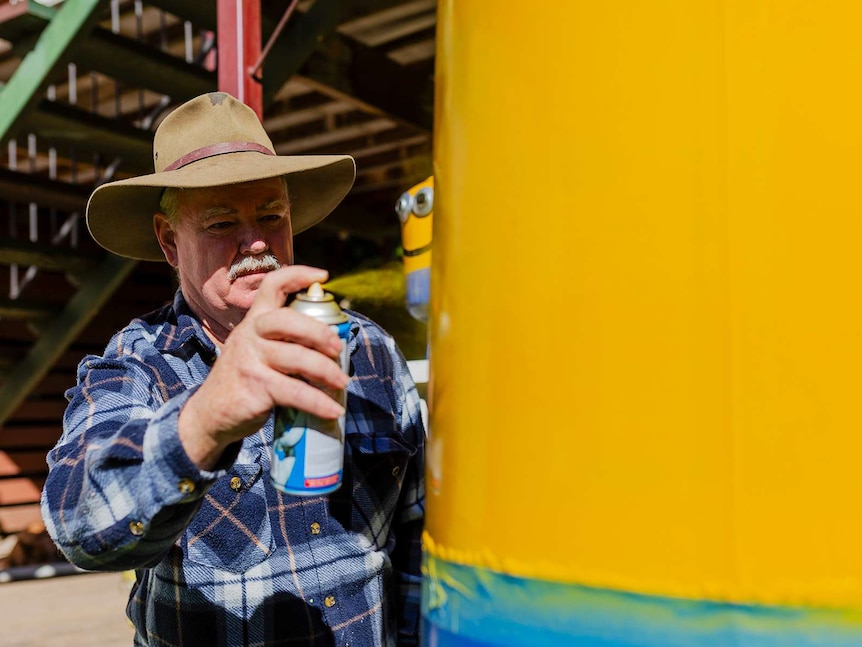 A man with an Akubra hat, spray paints a yellow drum (takes up half of right of photo). Small Minion visible in background.