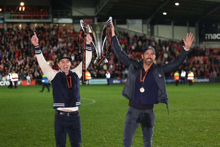 Wrexham owners Rob McElhenney and Ryan Reynolds hold up a trophy on the field after winning the National League.