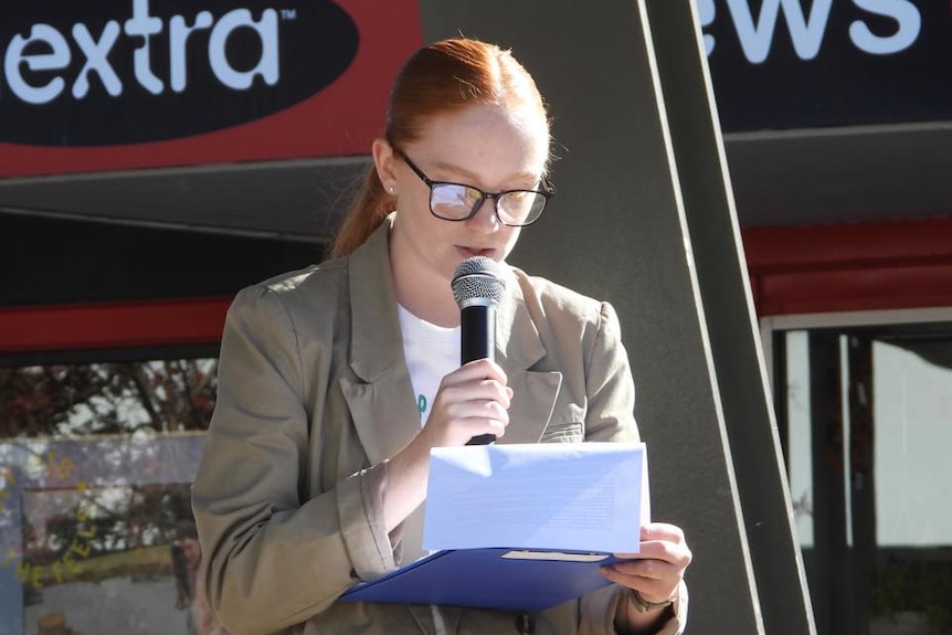 A young woman with red hair and a formal jacket makes a speech.