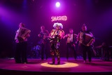 A group of five people play the accordian on stage under a neon sign that says 'smashed'.