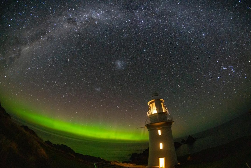 A green-tinged aurora australias behind a lit-up lighthouse