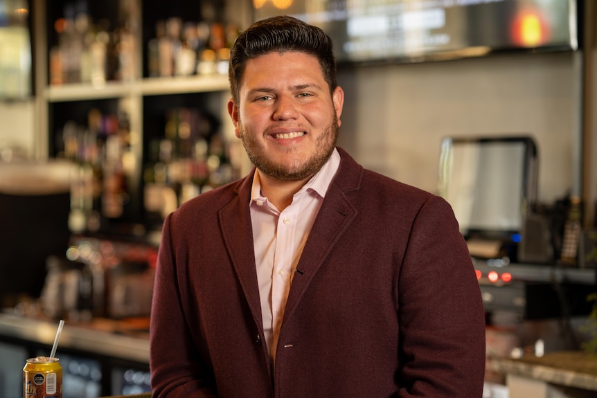 A man in a brown suit jacket with a beard stands in front of a bar