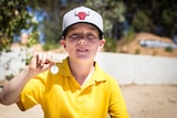 A nine-year-old boy in a yellow shirt holds up a small badge reading "Ambassador 2016".