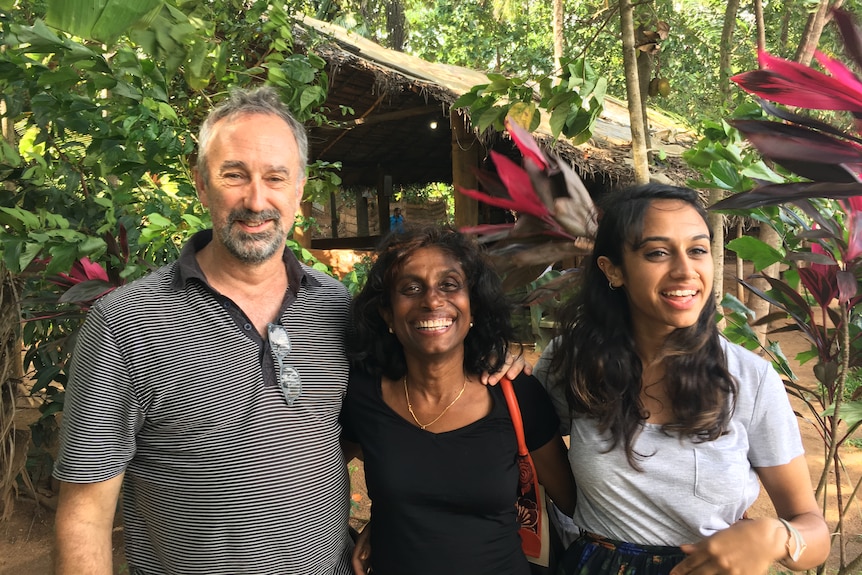 A man and woman stand arm in arm with their adult daughter. All are smiling.