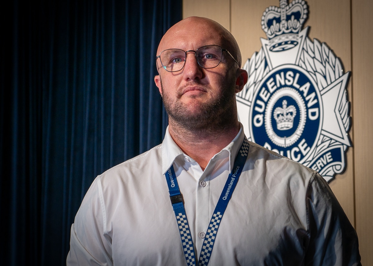 Police detective standing in front of Queensland Police logo and blue curtain