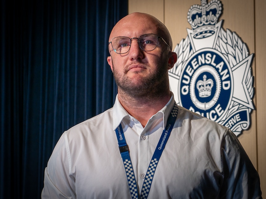 Police detective standing in front of Queensland Police logo and blue curtain