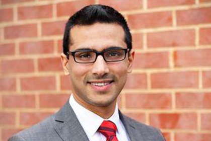 A man in glasses and red tie and suit standing against a brick background smiles to the camera.
