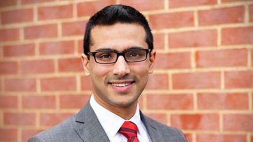 A man in glasses and red tie and suit standing against a brick background smiles to the camera.