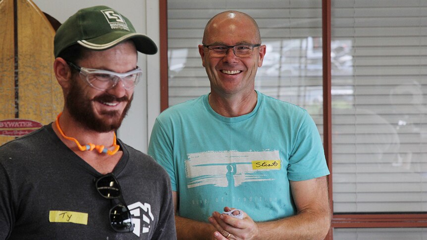Two men smiling and laughing in a workshop with a surf board in the background.