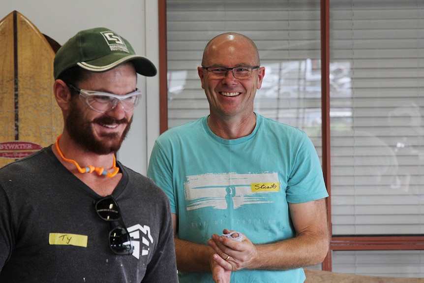 Two men smiling and laughing in a workshop with a surf board in the background.