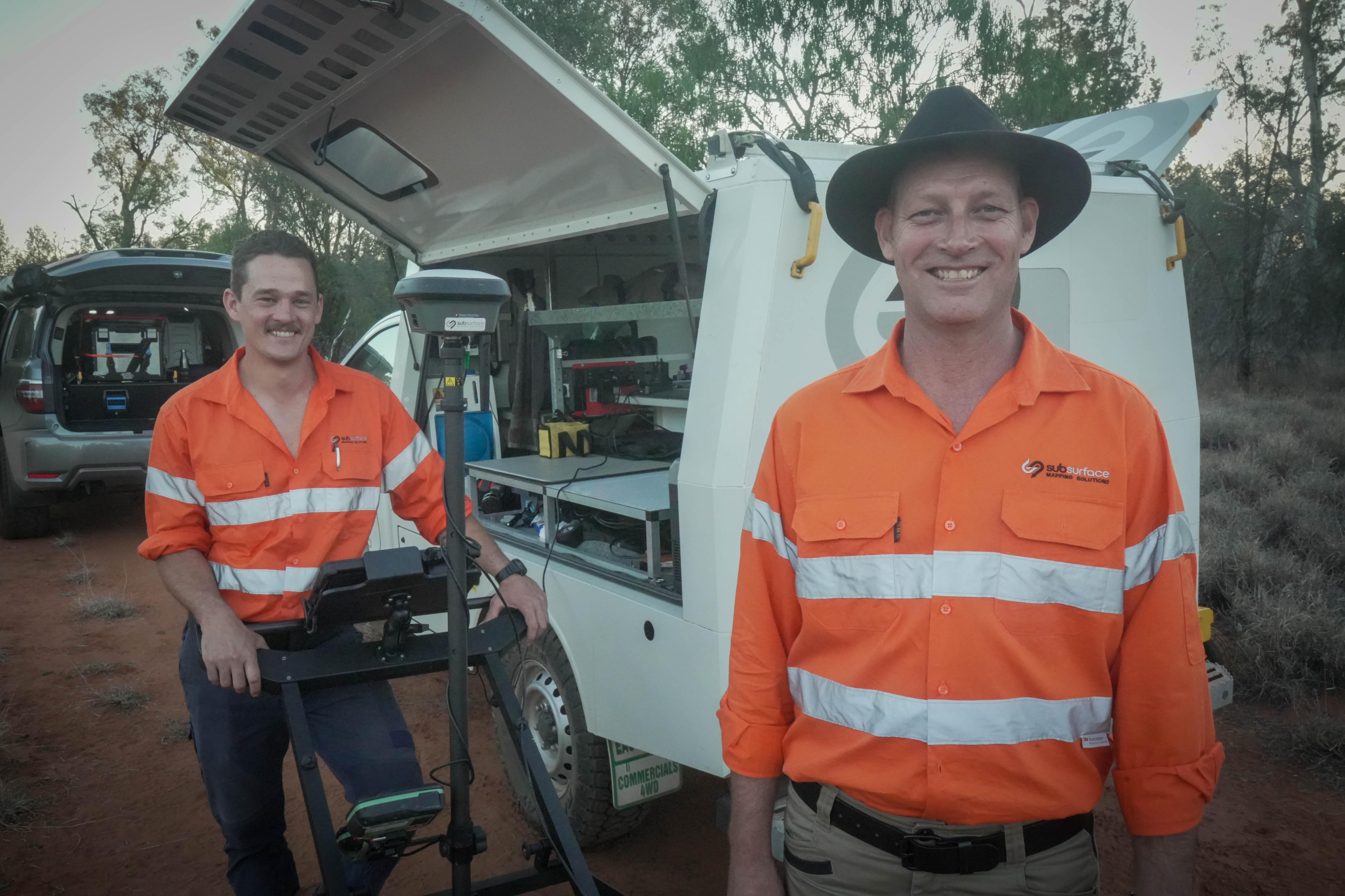 two men wearing high-vis orange shirts stand outside a van filled with technology.