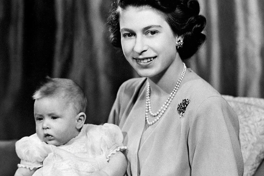 Queen Elizabeth II wearing a pearl necklace and blouse sits while holding a baby Prince Charles dressed in white