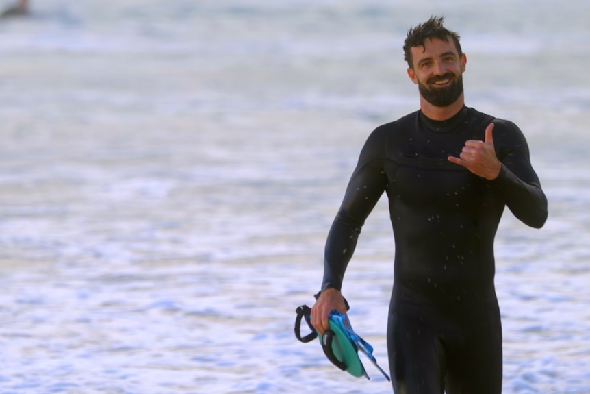 Man in wetsuit walking along the beach, surfer in background. 