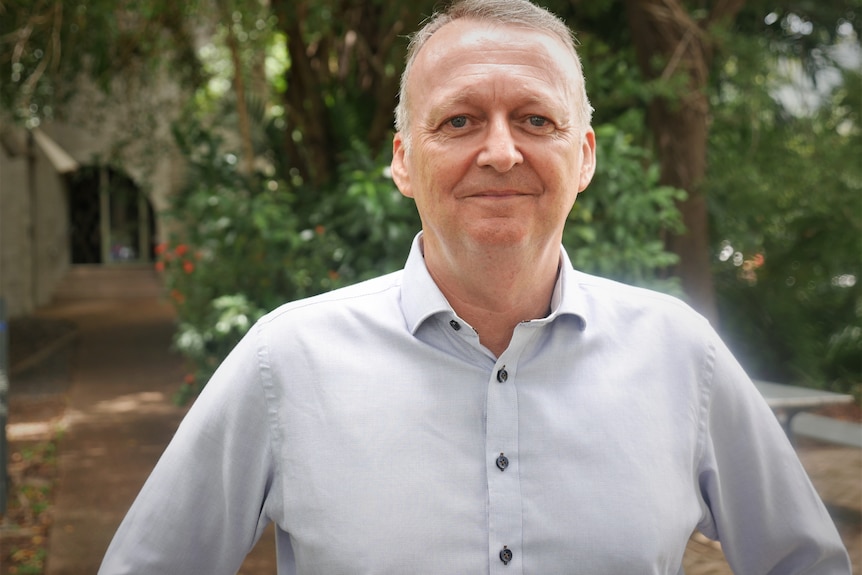 A man in a light blue shirt stands in front a bush smiling.