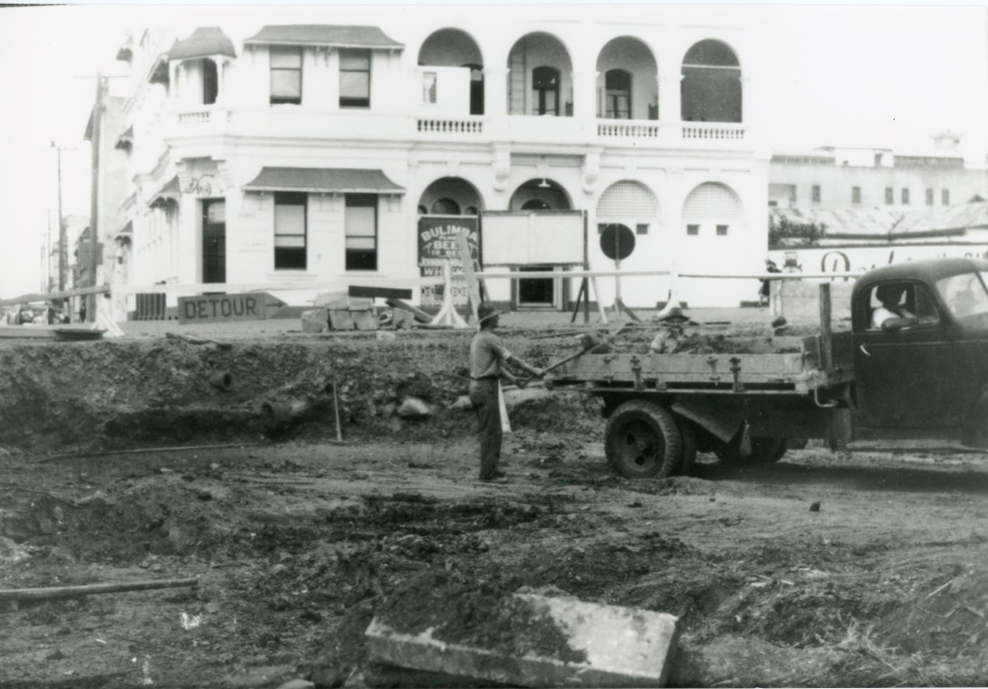 Black and white image of dirt dug up, man with pick up truck, two storey hotel building in background
