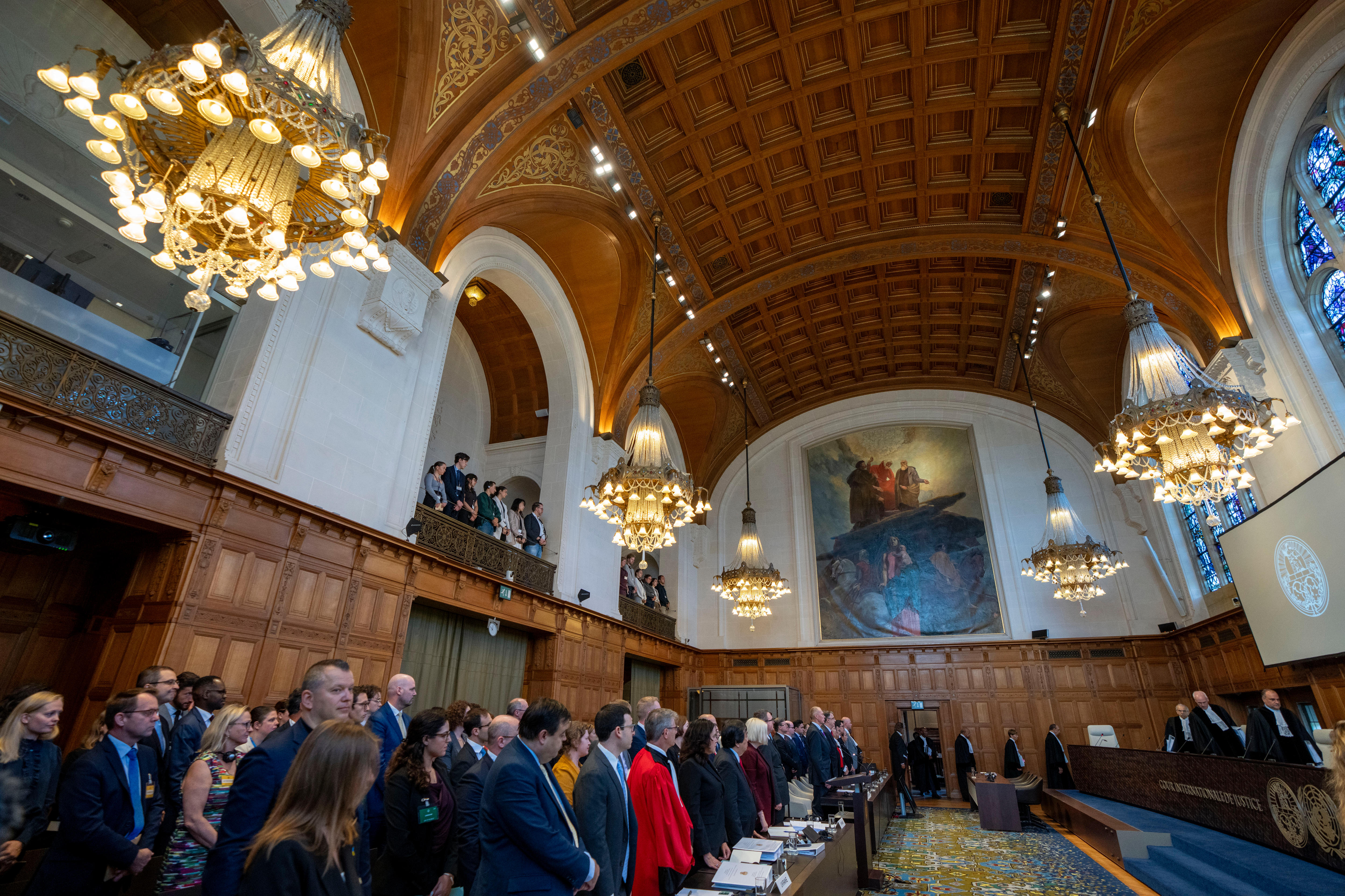 A crowd of people stand inside the World Court, which has high ceilings and large paintings on the walls.