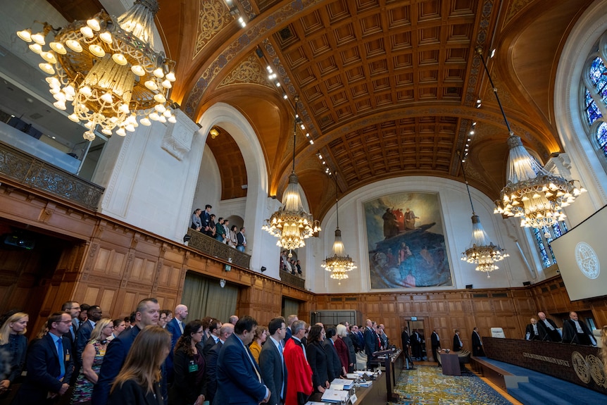 A crowd of people stand inside the World Court, which has high ceilings and large paintings on the walls.