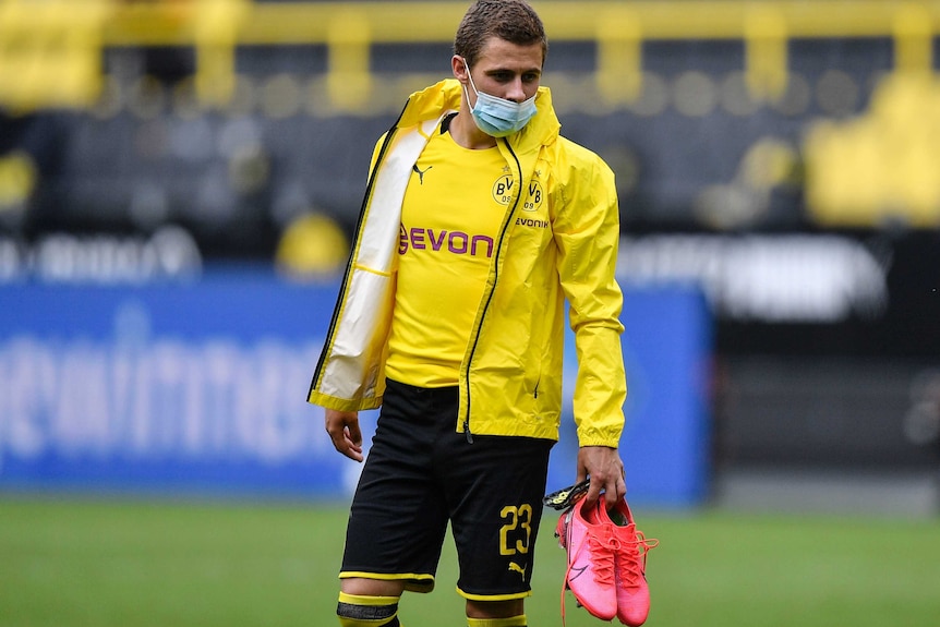 Thorgan Hazard wears a facemask and a yellow football kit, holding some red boots in his left hand