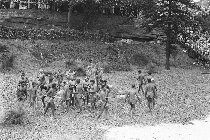 Black and white image of a group of aboriginal men wearing tradition dress, dancing in a circle.