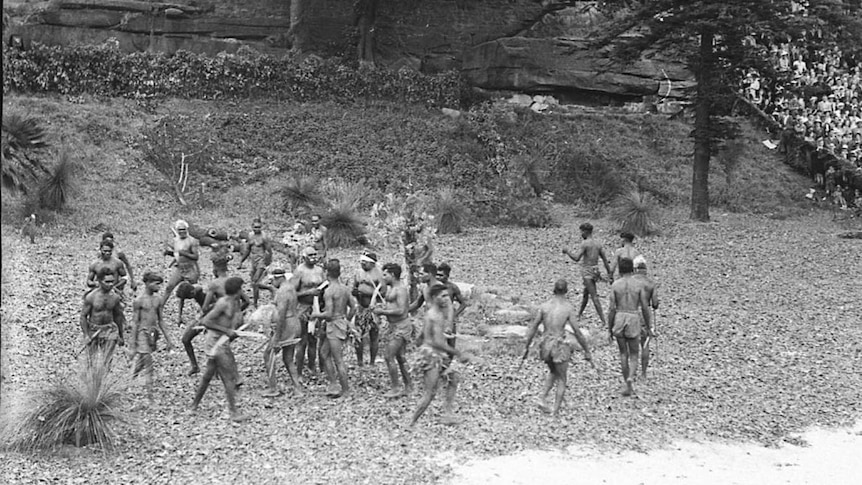 Black and white image of a group of aboriginal men wearing tradition dress, dancing in a circle.