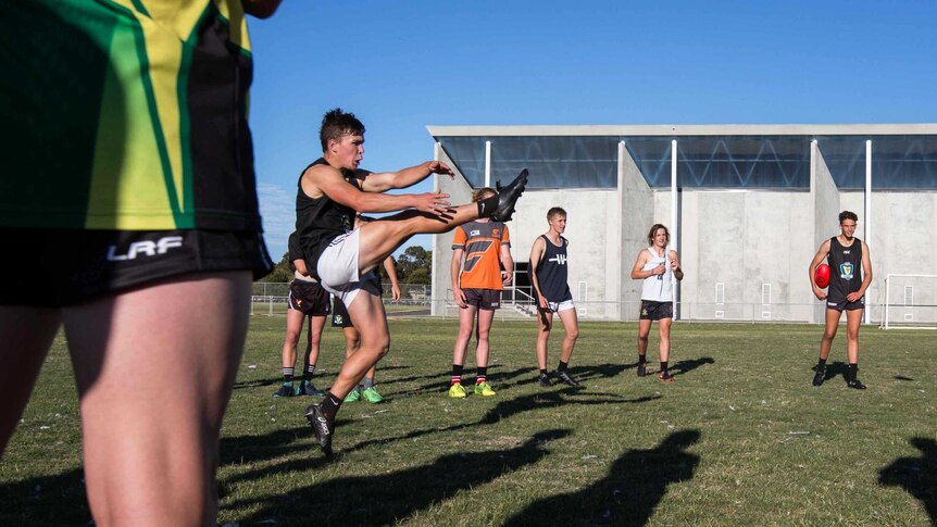 Footballer kicks ball at training, watched by teammates, Tasmania.