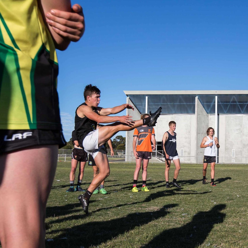 Footballer kicks ball at training, watched by teammates, Tasmania.