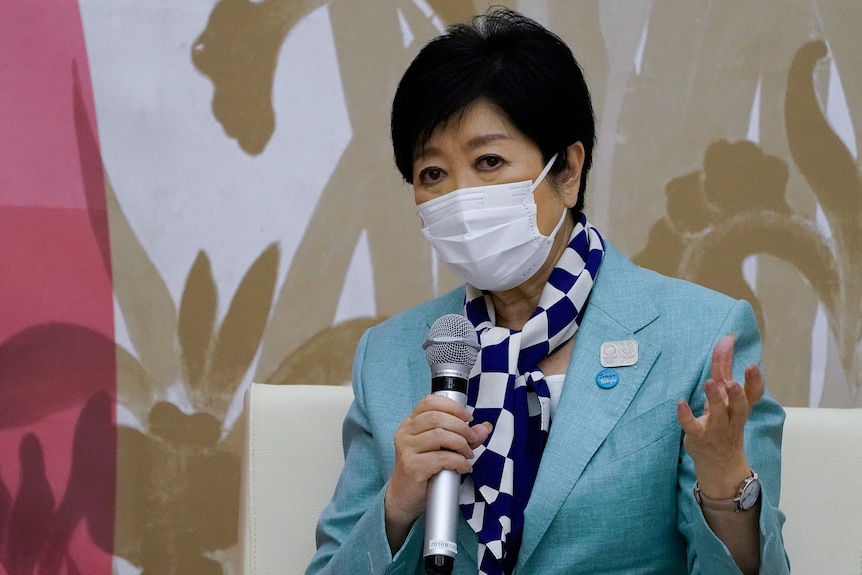 Yuriko Koike in a mask and blue suit with checkered scarf holds a microphone to speak. 