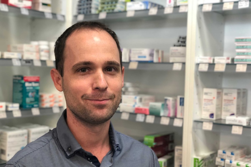 A man looking at the camera with pharmaceuticals on shelves behind him