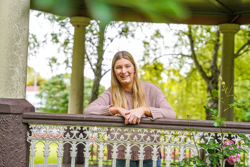 A teenage girl with blonde hair wearing a knitted jumper leans against a rotunda rail laughing.