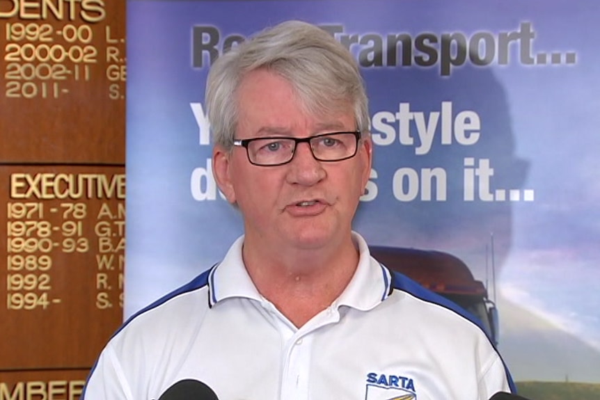 A man with grey hair and black glasses in a white shirt stands in front of a blue banner. 