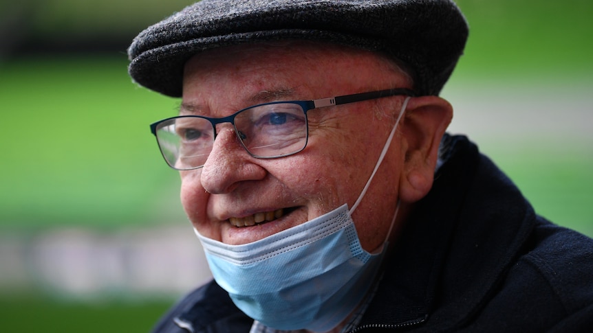 An elderly man wearing a cap and a facemask on his chin