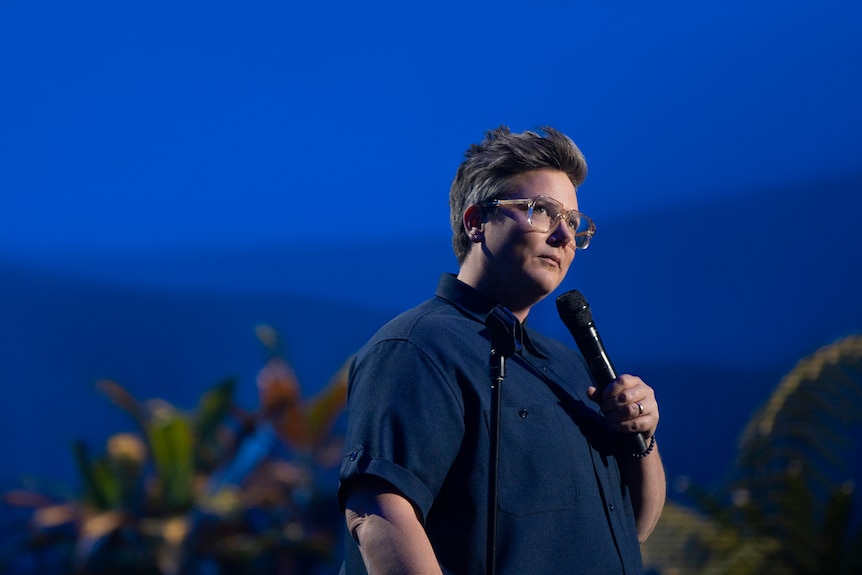Comedian Hannah Gadsby, with short hair, glasses and a dark blue shirt, holds a microphone on-stage.