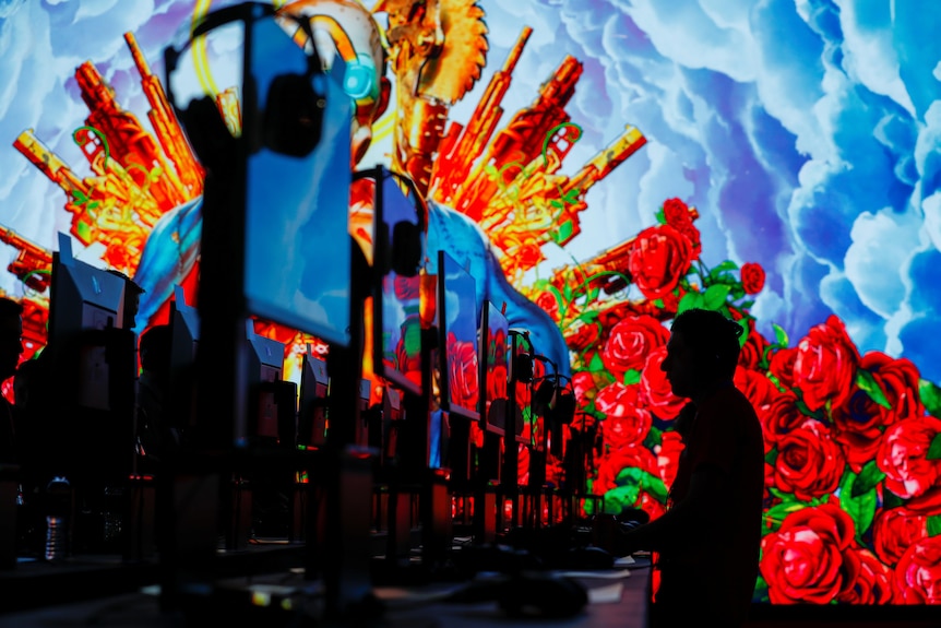 A row of computrers with headphones and one person playing with bright coloured clouds and roses in the background. 