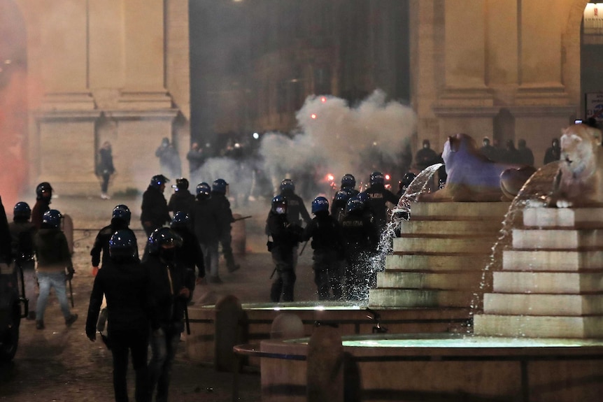 Riot police patrol with shields and batons as smoke billows during an evening  protest.