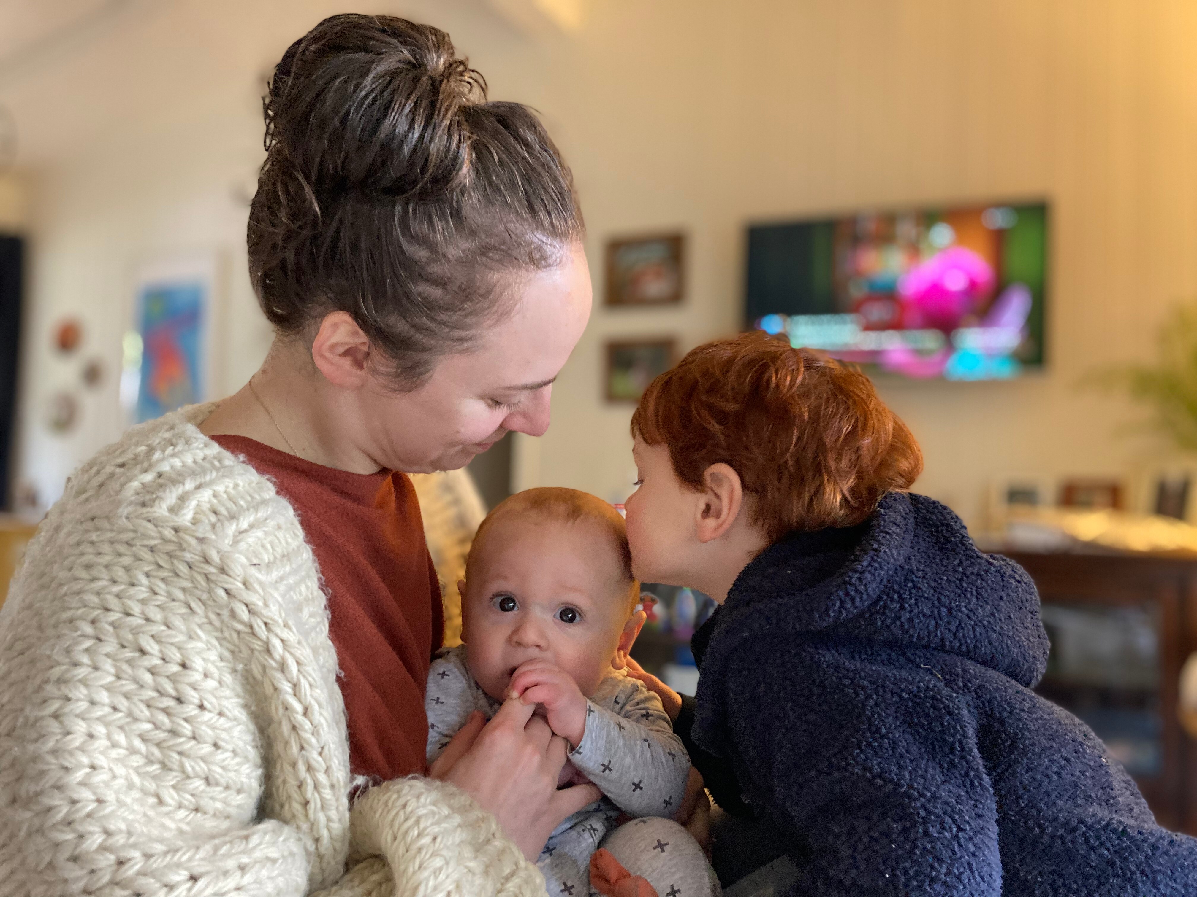 A woman sits with her baby in her living room as her young son gives the baby a kiss.